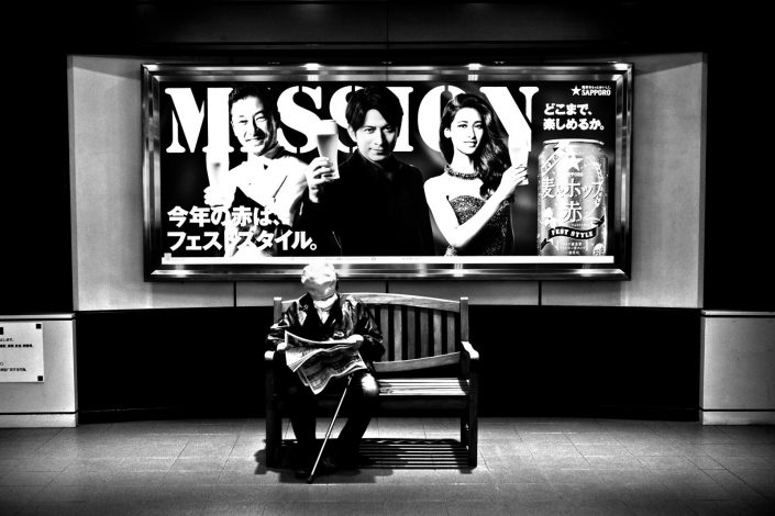 Old Japanese man sitting and reading a newspaper under a flashy billboard. Street photography by Victor Borst