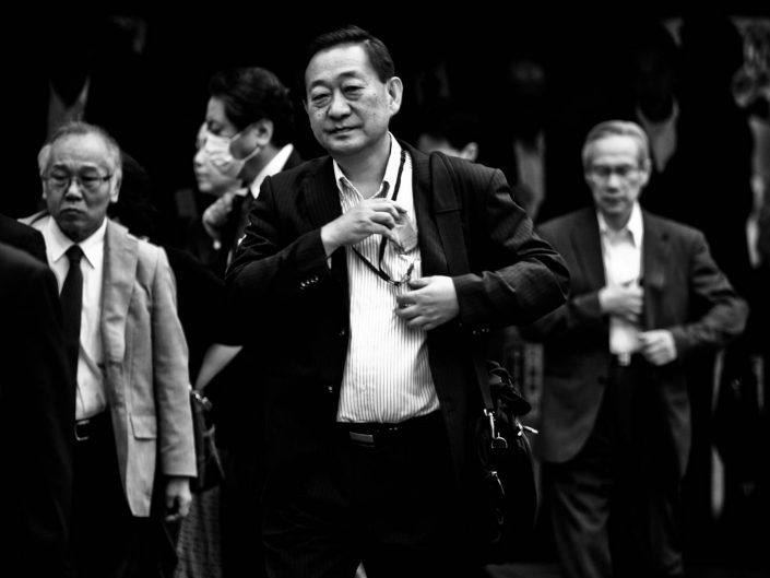 Group of salarymen at Shimbashi station with on the foreground one smiling. Street Photography by Victor Borst