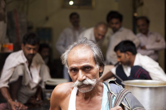 Colorful India, Indian man in the forefront with a mustache and people in dof cooking stuff. Made in Varanasi.